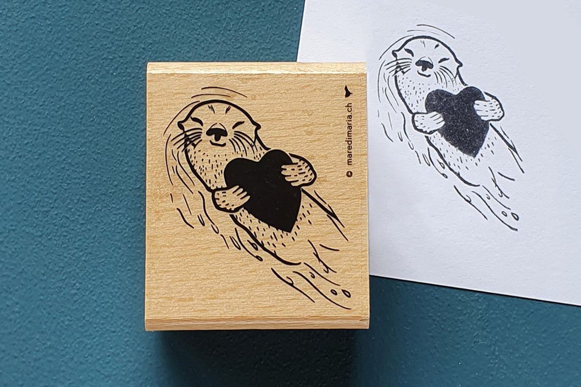 Holzstempel «Otterliebe» von Mare di Maria – rechteckiger Holzstempel mit Otter, der auf dem Rücken treibt und ein Herz hält, auf dunkelgrünem Grund, daneben der schwarze Abdruck auf weissem Papier