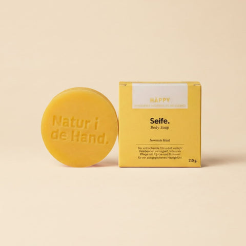 Savon naturel Häppy de Natur i de Hand – savon rond jaune avec le logo Natur i de Hand en relief, à côté de sa boîte jaune