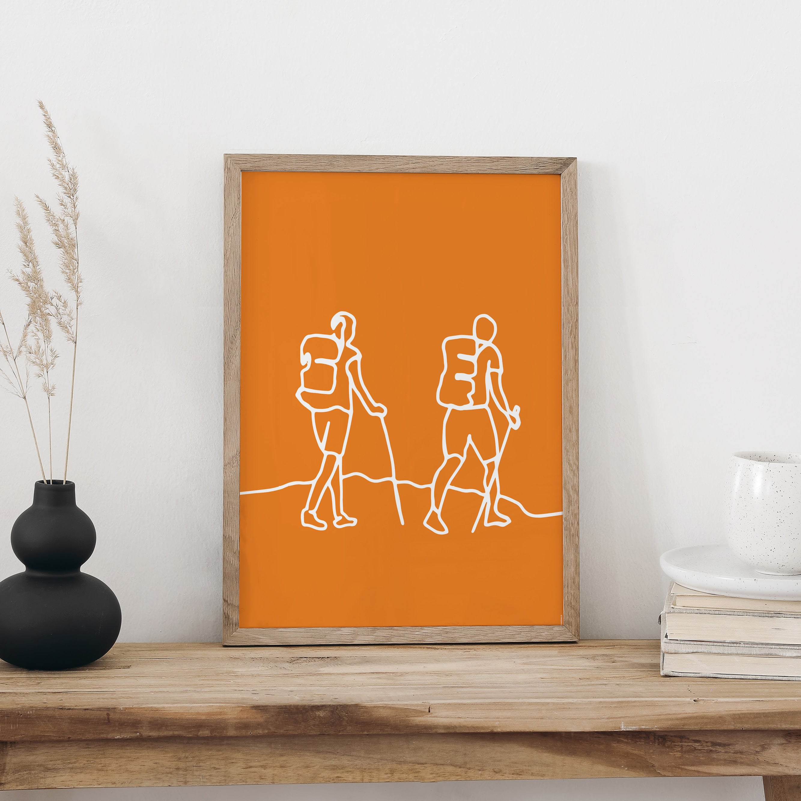 Plakat «Wandern» im hellen Holzrahmen auf einem Holztisch an weisser Wand, daneben eine schwarze Vase mit Trockengräsern und ein Bücherstapel mit Tasse