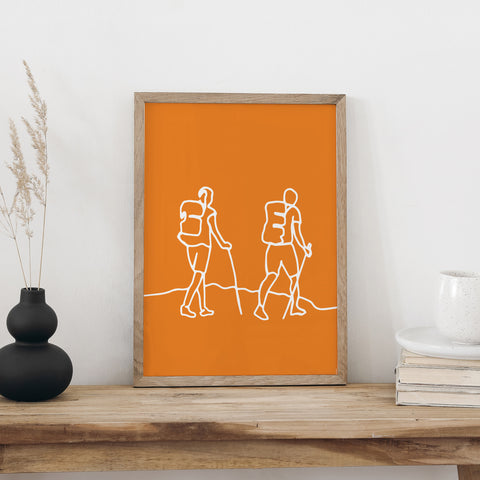 Plakat «Wandern» im hellen Holzrahmen auf einem Holztisch an weisser Wand, daneben eine schwarze Vase mit Trockengräsern und ein Bücherstapel mit Tasse