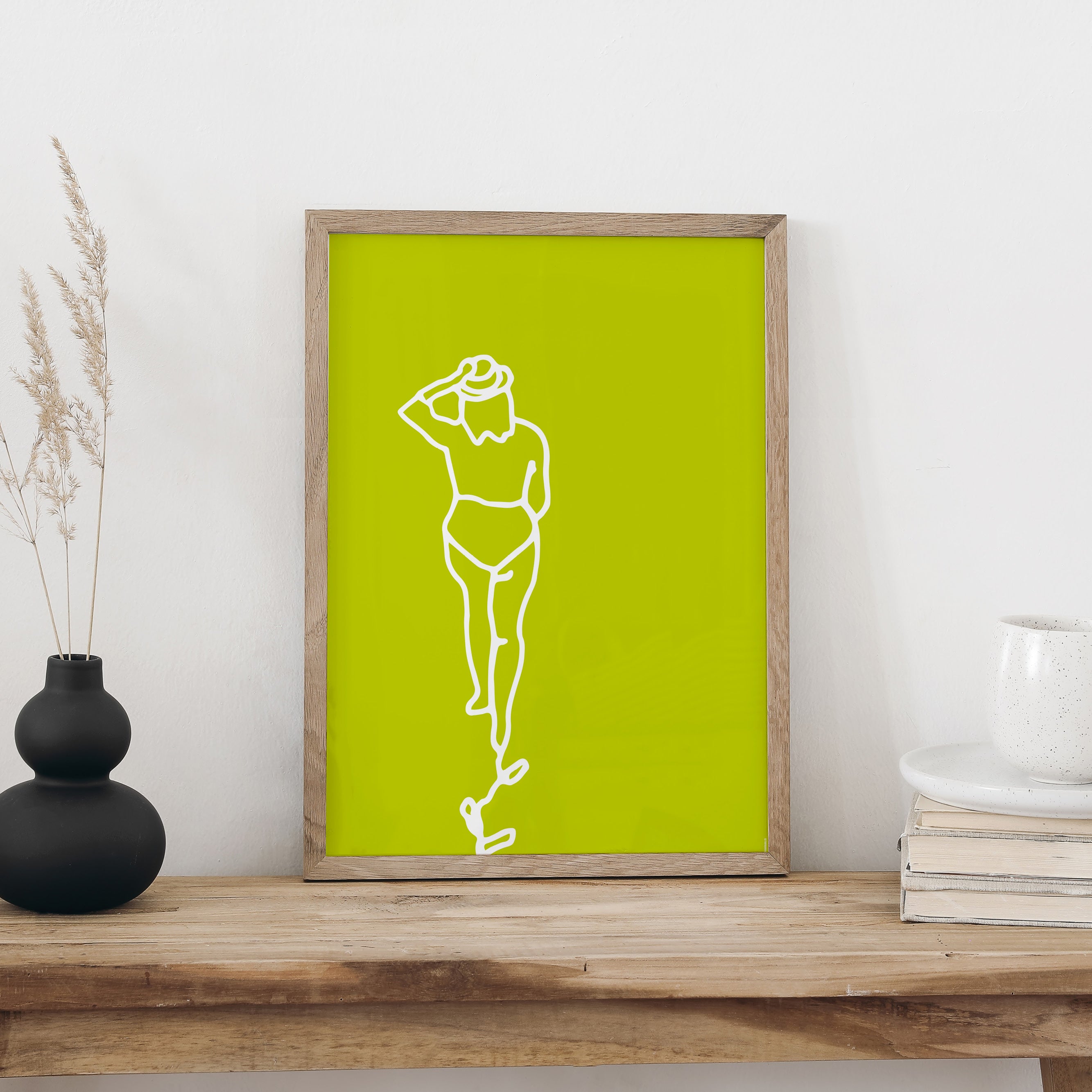 Plakat «Strandspaziergang» im hellen Holzrahmen auf einem Holztisch an weisser Wand, daneben eine schwarze Vase mit Trockengräsern und ein Bücherstapel mit Tasse