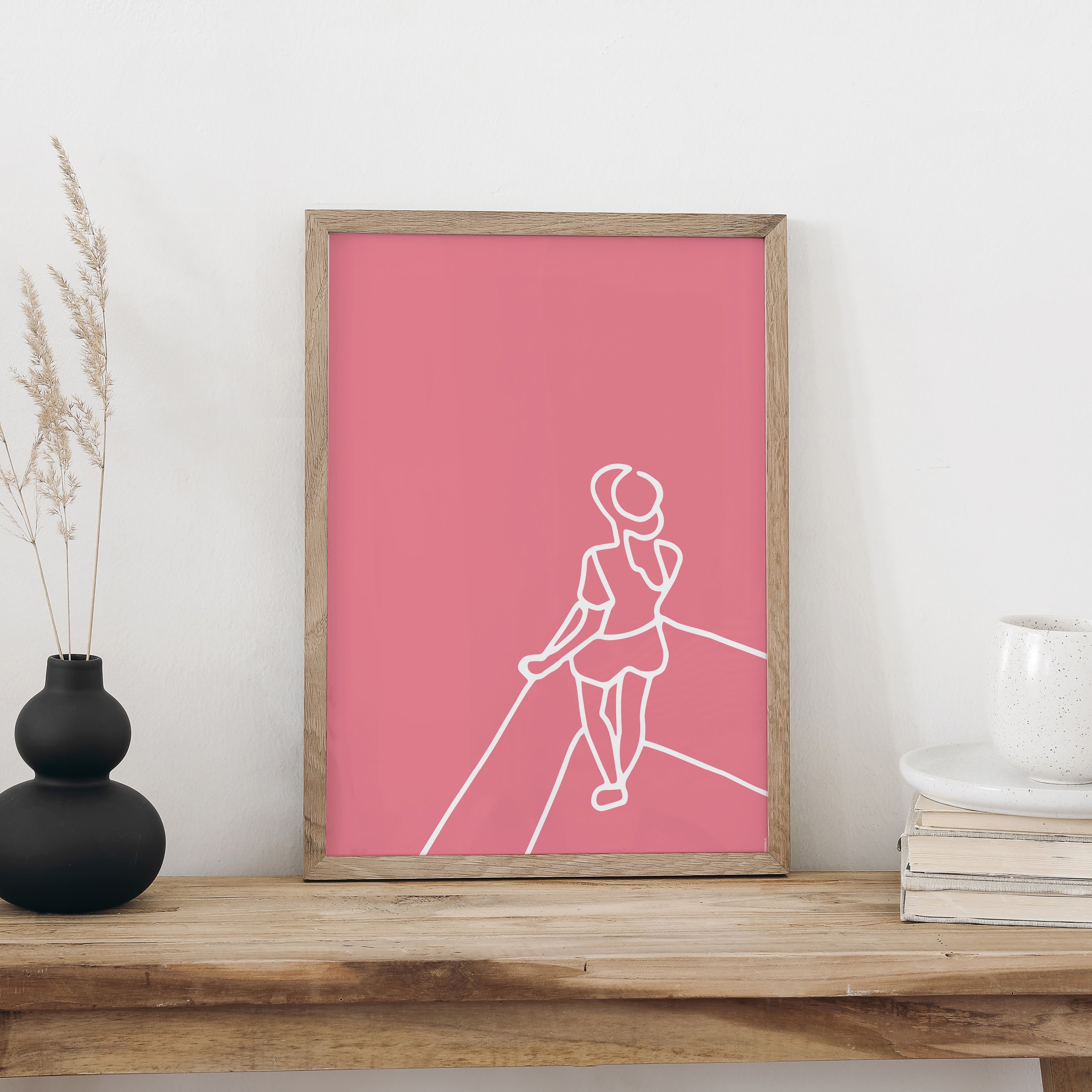 Plakat «Aussicht» im hellen Holzrahmen auf einem Holztisch an weisser Wand, daneben eine schwarze Vase mit Trockengräsern und ein Bücherstapel mit Tasse