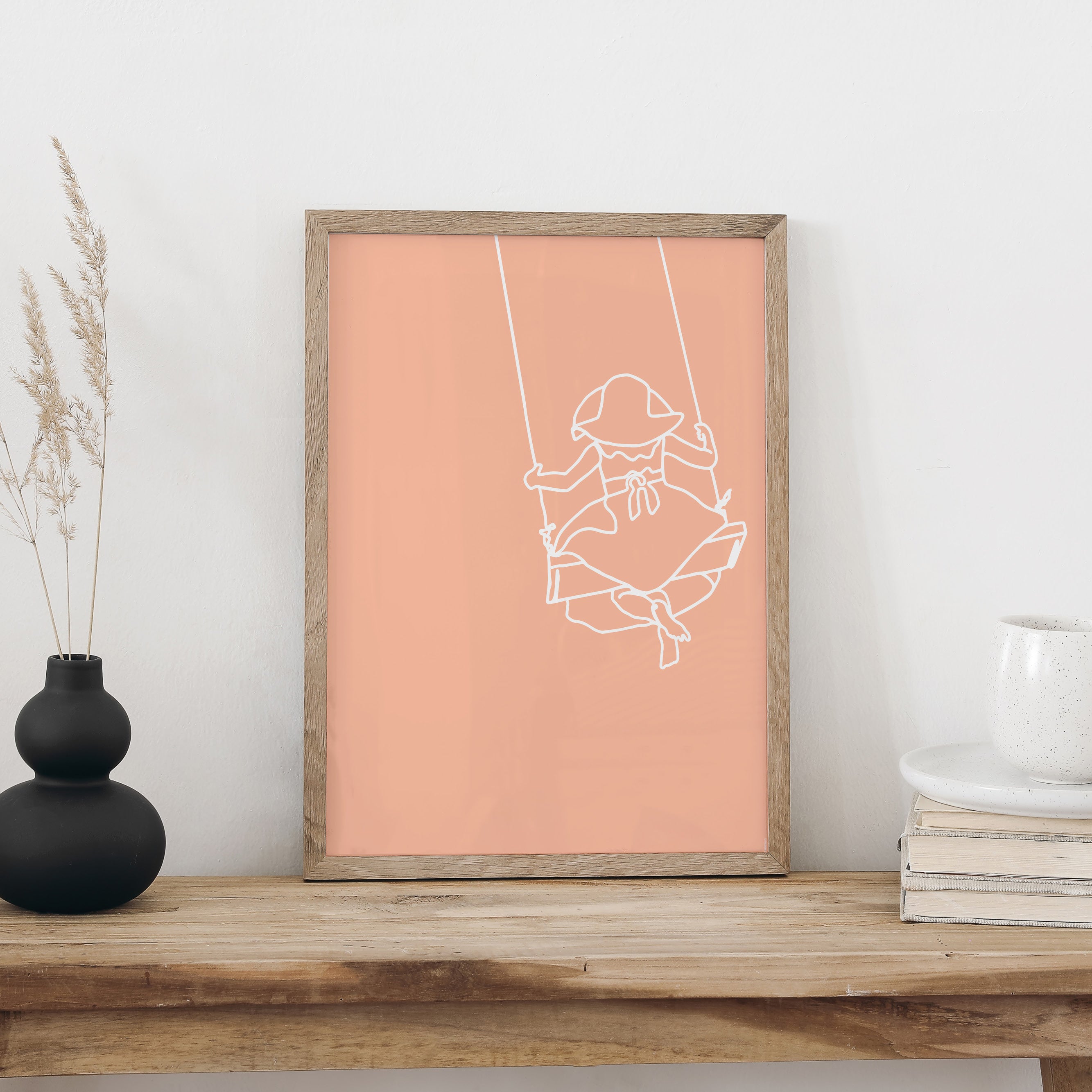 Plakat «Schaukeln» im Holzrahmen auf einer Holzbank an weisser Wand, daneben eine schwarze Vase mit Trockengräsern und ein Bücherstapel mit Tasse