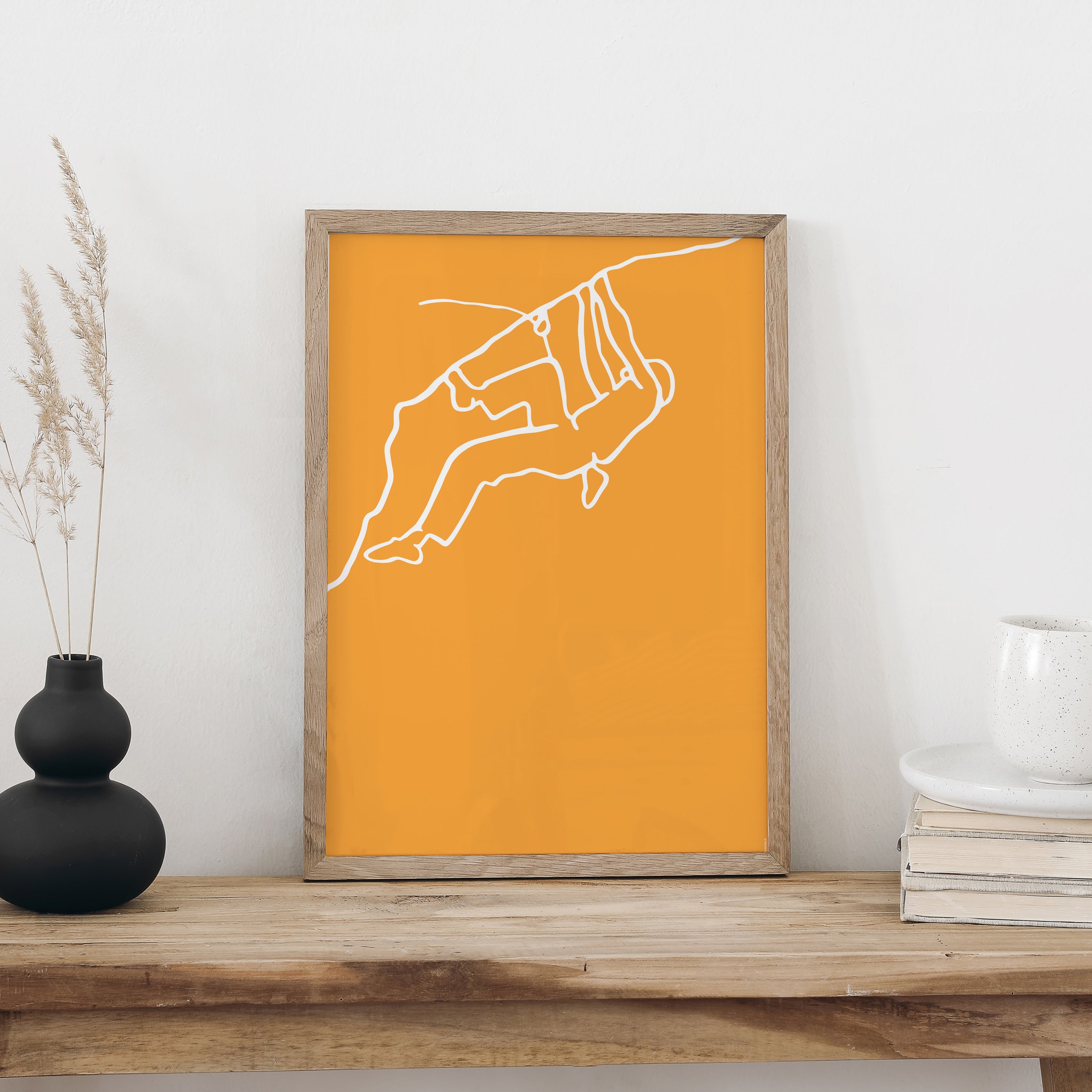 NaniManu Plakat «Klettern» im Holzrahmen auf einer Holzbank an weisser Wand, daneben schwarze Vase mit Trockengräsern und ein Bücherstapel mit Tasse