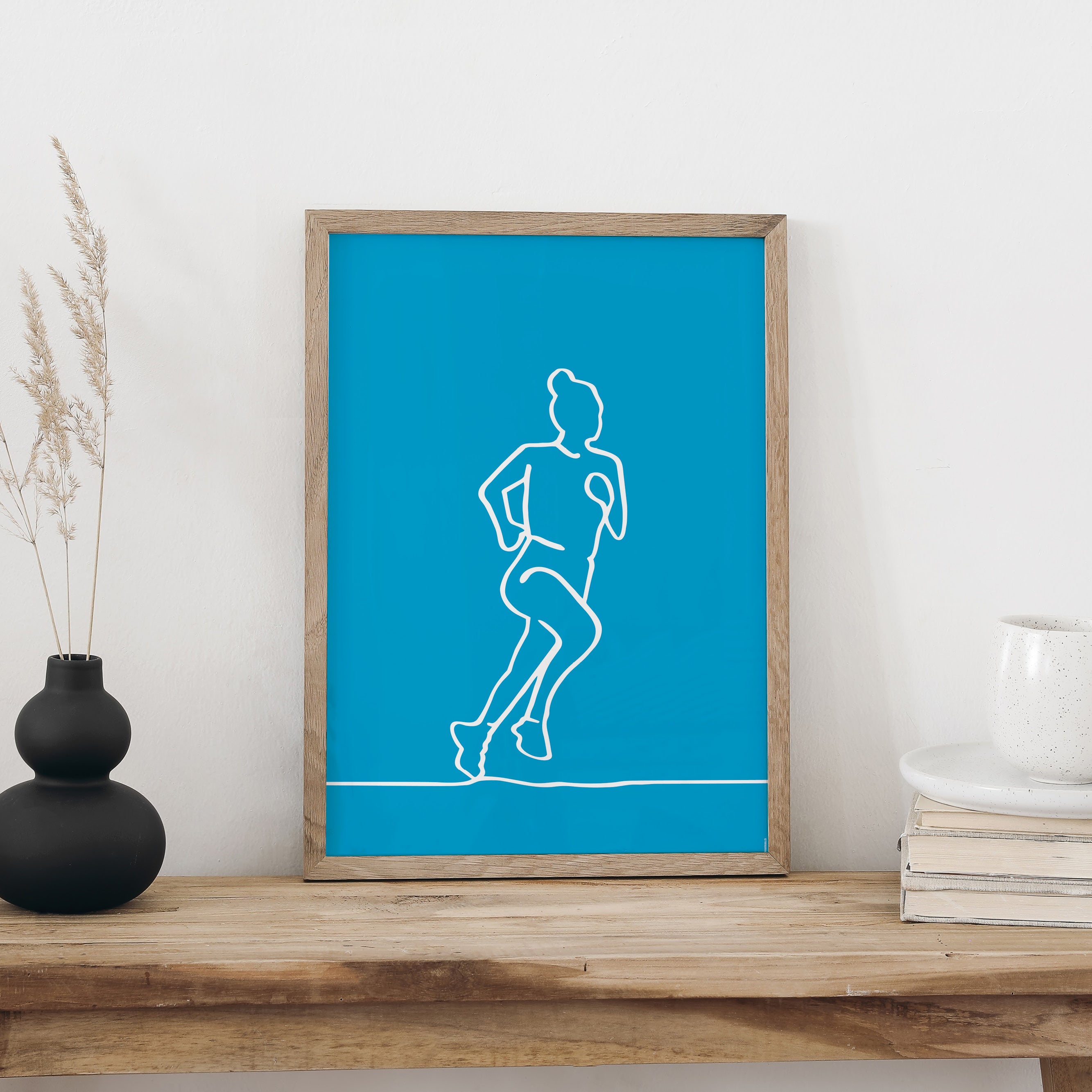 NaniManu Plakat «Rennen» im Holzrahmen auf einer Holzbank an weisser Wand, daneben schwarze Vase mit Trockengräsern und ein Bücherstapel mit Tasse