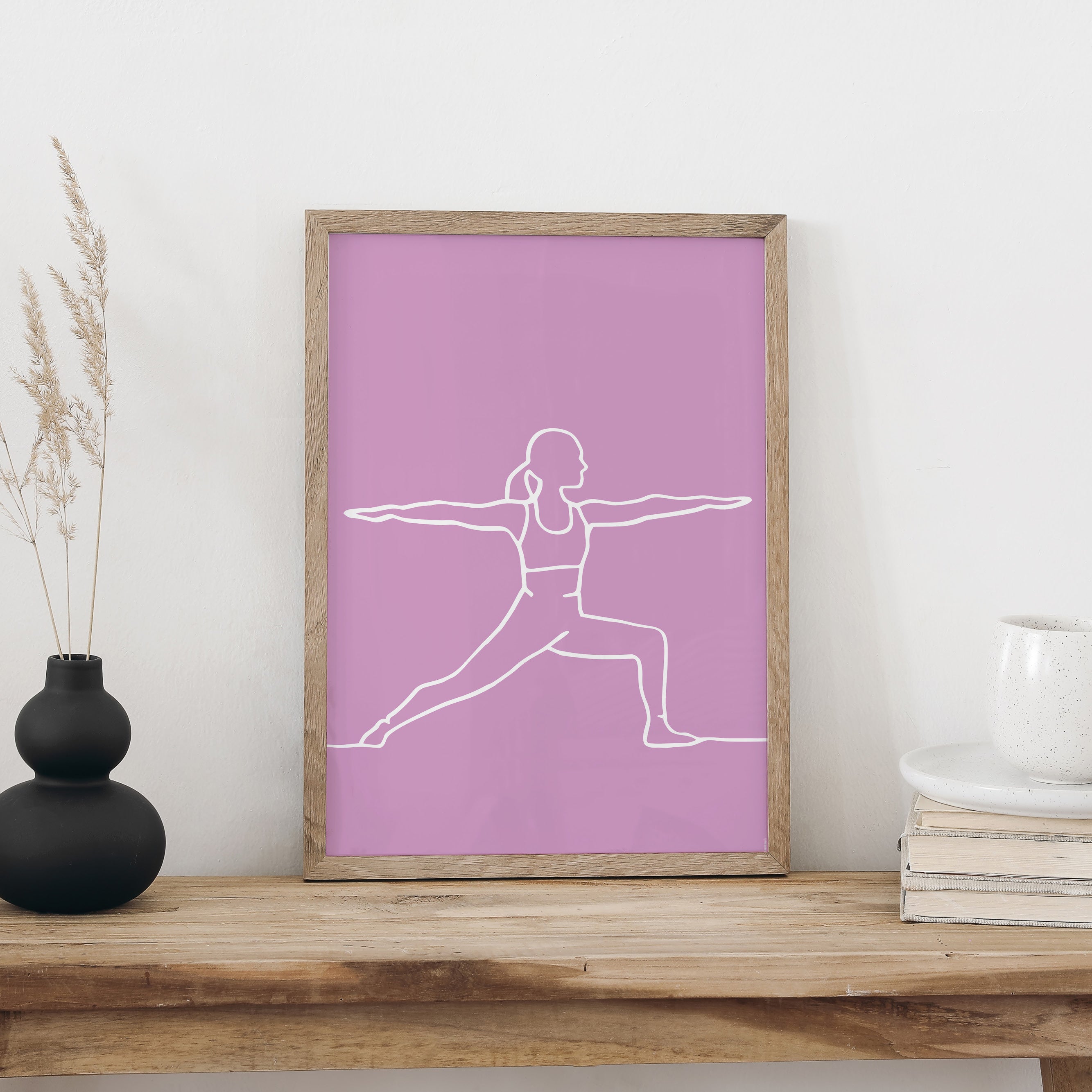 NaniManu Plakat «Yoga» im Holzrahmen auf einer Holzbank an weisser Wand, daneben schwarze Vase mit Trockengräsern und ein Bücherstapel mit Tasse