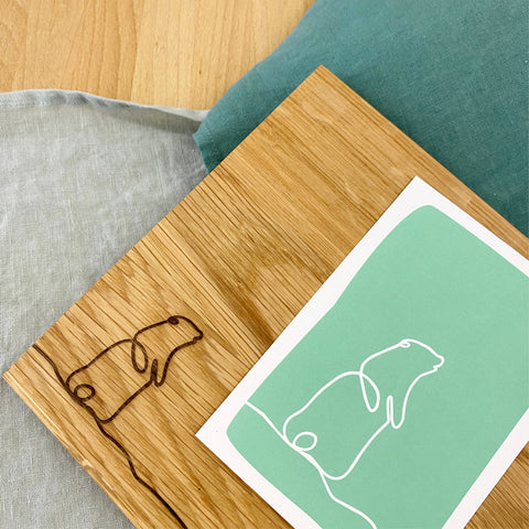 Detailaufnahme der Murmeltier-Gravur auf Eiche Grösse S, daneben eine Postkarte mit demselben Strichmotiv auf grünem Grund