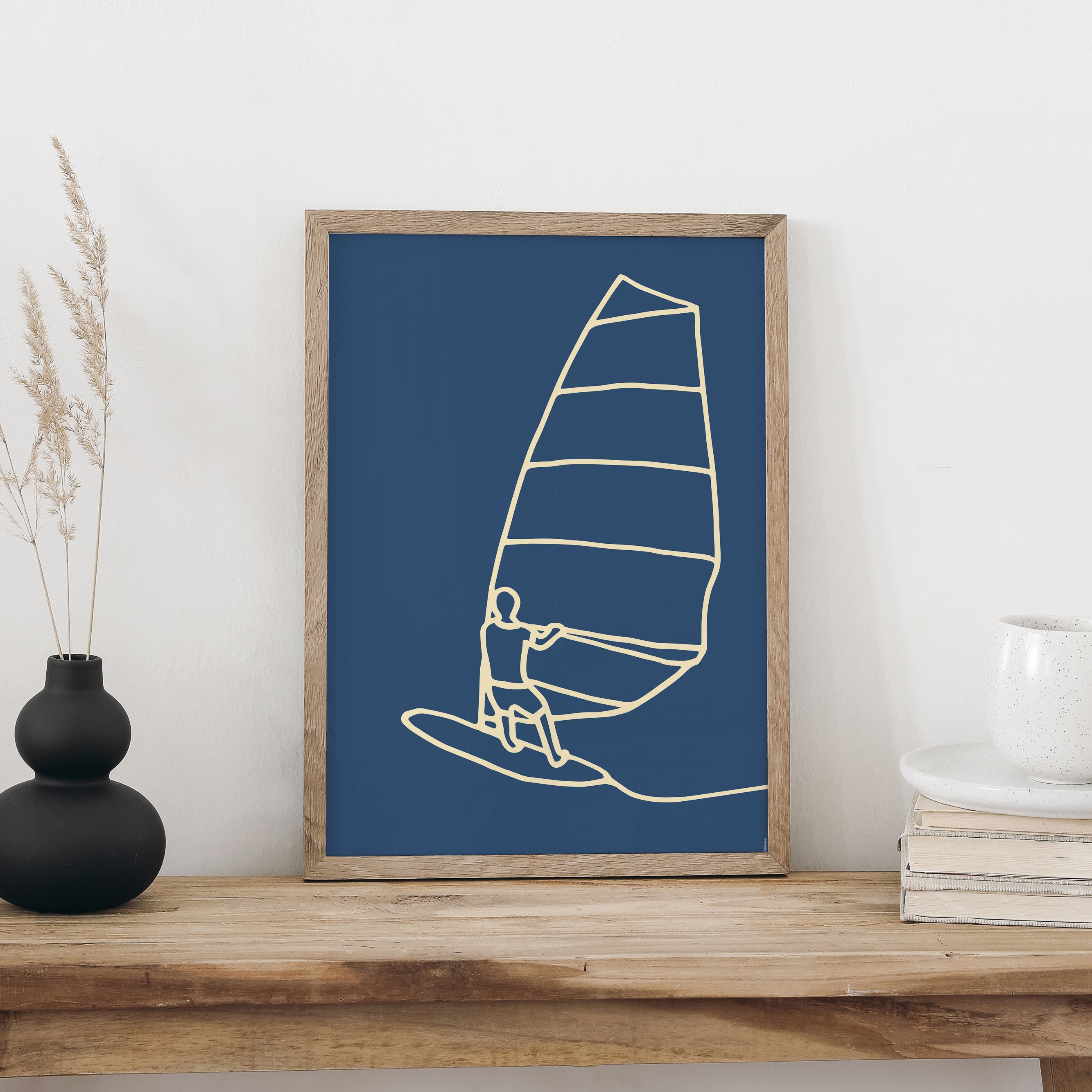 Windsurfen-Plakat im Holzrahmen auf einer Holzbank, daneben eine schwarze Vase mit Trockengräsern und eine weisse Tasse auf Büchern