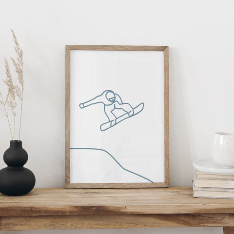 Snowboarden-Plakat im Holzrahmen auf einer Holzbank, daneben eine schwarze Vase mit Trockengräsern und eine weisse Tasse auf Büchern