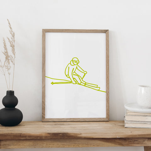 Skifahren-Plakat von NaniManu im Holzrahmen auf einer Holzbank, daneben eine schwarze Vase mit Trockengräsern und ein Bücherstapel mit Tasse