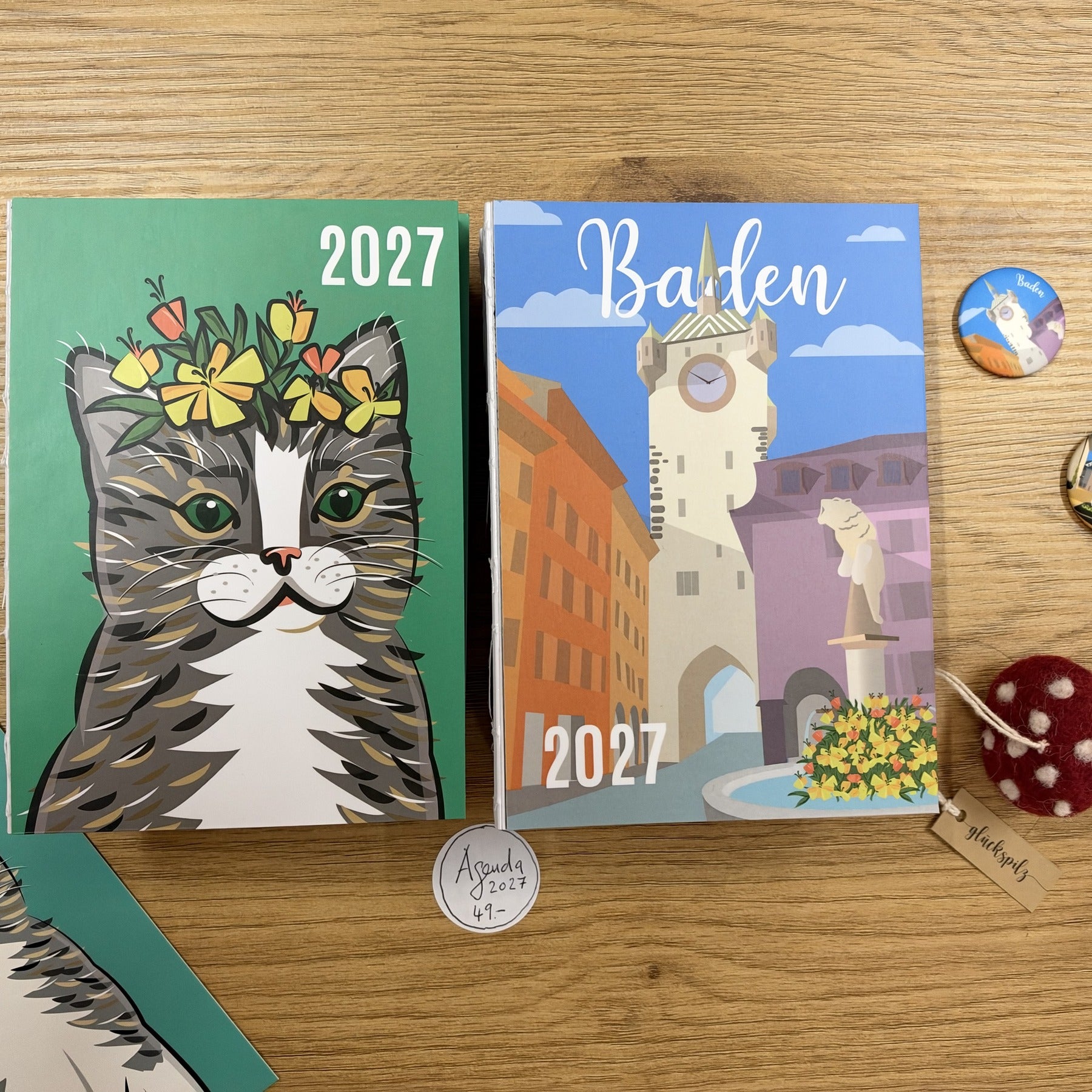 The 2027 agendas Cat Sir James and Stadtturm Baden on a wooden table, next to Baden badges and a lucky mushroom