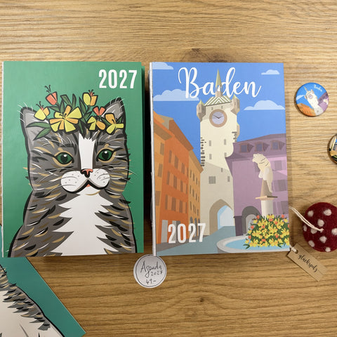 The 2027 agendas Cat Sir James and Stadtturm Baden on a wooden table, next to Baden badges and a lucky mushroom