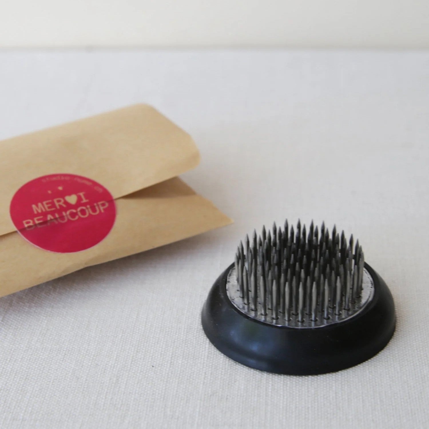 Kenzan flower frog with metal pins in a black base, next to a paper bag with a red «merci beaucoup» sticker