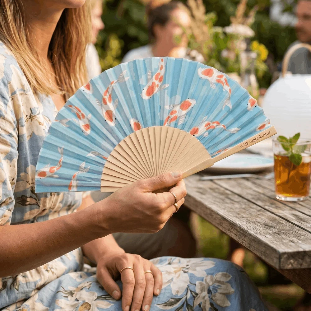 Der Koi-Fächer in der Hand einer Frau an einem sonnigen Gartentisch, im Hintergrund Gäste und ein Glas Eistee