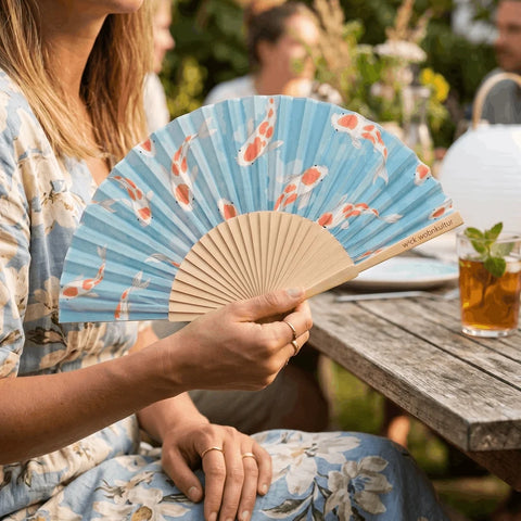 Der Koi-Fächer in der Hand einer Frau an einem sonnigen Gartentisch, im Hintergrund Gäste und ein Glas Eistee