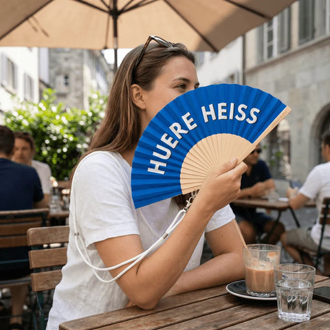 An einem Cafétisch in einer Altstadtgasse hält eine Frau den Fächer «Huere Heiss» vor ihr Gesicht