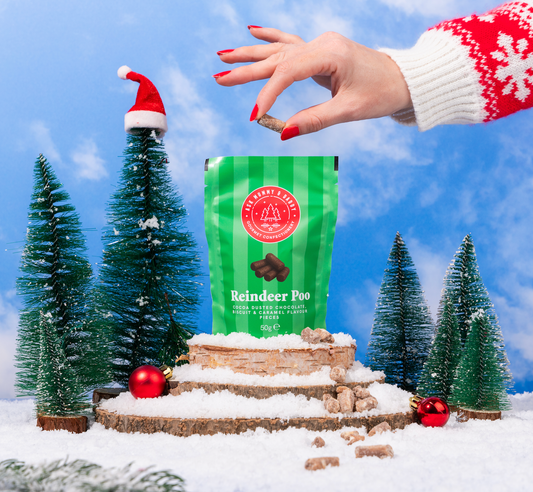 NaniManu Reindeer Poo (Ask Mummy & Daddy) – grüner Beutel in einer verschneiten Winterszene, eine Hand nimmt ein Schoggi-Stückchen