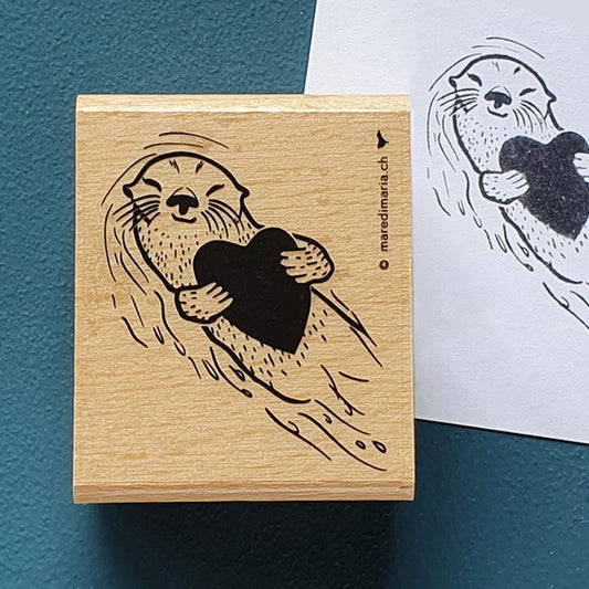 Holzstempel «Otterliebe» von Mare di Maria – rechteckiger Holzstempel mit Otter, der auf dem Rücken treibt und ein Herz hält, auf dunkelgrünem Grund, daneben der schwarze Abdruck auf weissem Papier